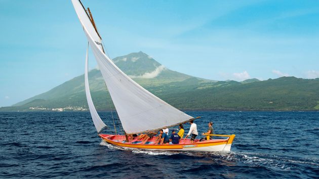 A yacht at a slight angle, sailing through water. The island's mountain is behind.