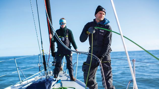 Two people in sailing gear on a yacht holding ropes.