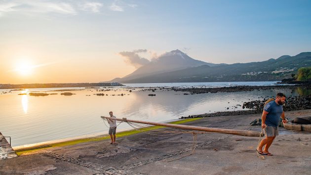 Two people carrying a mast on the beach with the mountain behind